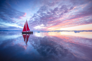 Sail boat with red sails cruising among ice bergs during sunrise. Disko Bay, Greenland.