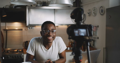 Happy young smart black blogger man filming new vlog video with professional camera in kitchen at...