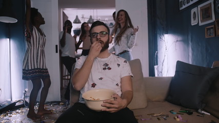 Young smart Caucasian man is sad and tired, watching movie eating snacks at house party while...