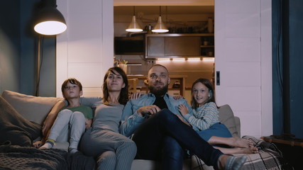 Happy young European family with two kids gather on couch to watch TV at home smiling. Togetherness and entertainment.