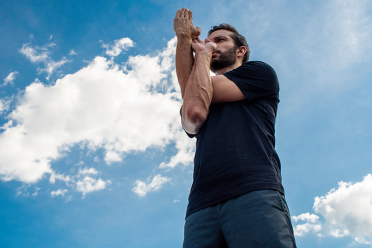 Man wearing a black shirt and dark green shorts stands straight up with his hands in eagle pose correcting his bodies posture in front of a bright blue sky with gorgeous white clouds behind his body.