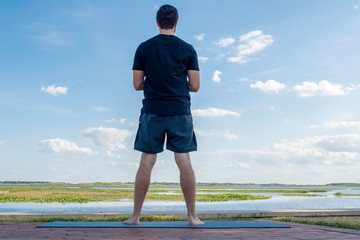 Man wearing a black shirt and dark green shorts stands looking outward over the sea in attempt to...