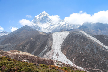 White snowy Ama Dablam mountain peak rises above mountain valley in Himalayas in the morning on the way to Everest base camp in Nepal. Clouds on the sky. Theme of beautiful mountain landscapes.