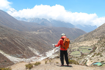 Person in orange windbreaker stands on mountainside during sunny day in Himalayas. Samdrang village is visible in valley in the background. Trek to Everest base camp. Theme of trekking in Nepal.