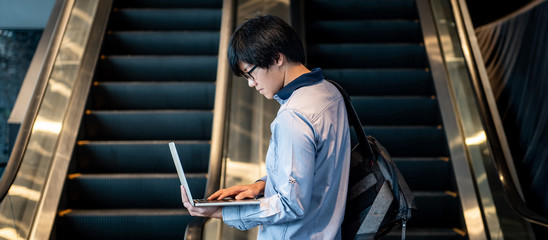 Asian man with glasses and bag holding laptop computer standing in front of escalator in public building. Working anywhere concept