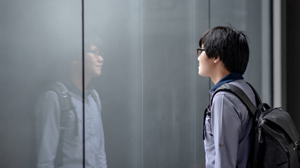 Young Asian man carrying bag looking himself on glass window while raining. Guy reflecting in the mirror. Emotional reflection concept