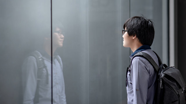 Young Asian man carrying bag looking himself on glass window while raining. Guy reflecting in the mirror. Emotional reflection concept