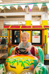 Obraz na płótnie Canvas A very joyful and happy little blond boy of three years old is riding in a yellow car with a green car in the summer Riviera Amusement Park, Sochi with his back to the viewer. Rest, tropics, rest