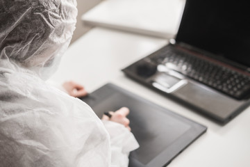 Girl in medical mask and white transparent protective suit sits in quarantine at home and works at computer and graphics tablet. Designer, artist, architect at remote work in a pandemic covid.