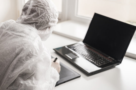 Girl in medical mask and white transparent protective suit sits in quarantine at home and works at computer and graphics tablet. Designer, artist, architect at remote work in a pandemic covid.