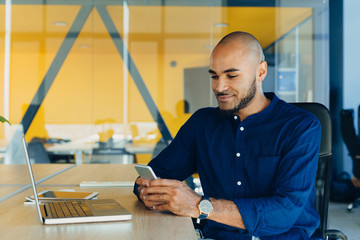 Handsome cheerful african american man in creative atmosphere using laptop sitting on a wooden table.