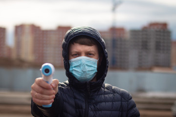 Young man in a dark jacket in a medical mask holds a non-contact thermometer on the street