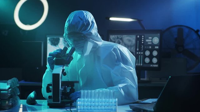 Scientist in protection suit and masks working in research lab using laboratory equipment: microscopes, test tubes. Coronavirus 2019-ncov hazard, pharmaceutical discovery, bacteriology and virology.