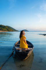 Beautiful brunette girl in a yellow dress sits in an old wooden boat overlooking a deserted tropical island. Romance at sea