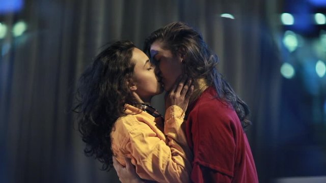 Girlfriends kissing each other at night in city with traffic motion passing in background. LGBT spontaneous kiss