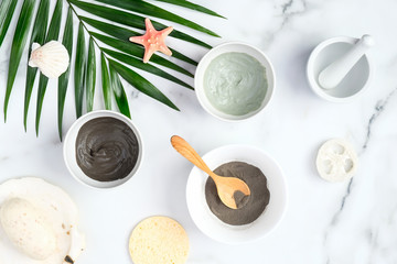 SPA natural organic facial clay mask and powder in bowls and tropical palm leaf on marble desk. Beauty products for face skin care and treatment concept.