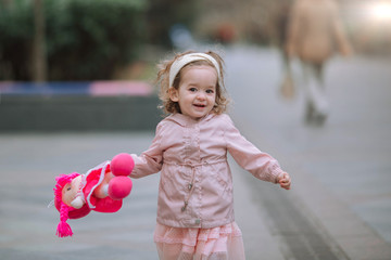 A nice little girl in a pink jacket and dress runs along the street and holds a doll in her hands.