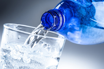 Pouring mineral water from blue bottle into clear glass on abstract grey background