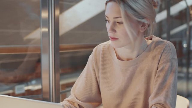Tilt up shot of young blonde businesswoman in casual outfit working on laptop and drinking tea while sitting at office desk