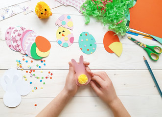 The child makes crafts with his own hands for the Easter. Colorful handmade from multi-colored paper. Scissors, cardboard, eggs, chicken, rabbit. Art creativity on a wooden table. Top view, copy space