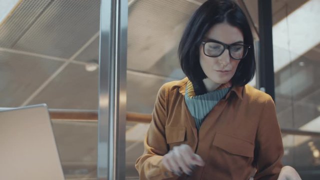 Caucasian brunette businesswoman in casual clothes and glasses typing on laptop and taking notes in notepad at desk in modern office with glass walls
