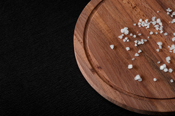 Wooden round cutting board on a black background. Empty board with crystals of salt closeup. 