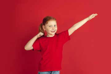 Winner gesture. Caucasian little girl portrait isolated on red studio background. Cute redhair model in red shirt. Concept of human emotions, facial expression, sales, ad, childhood. Copyspace.