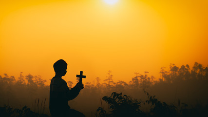 Human kneeling down with holding the cross and praying at sunset background. christian silhouette concept.