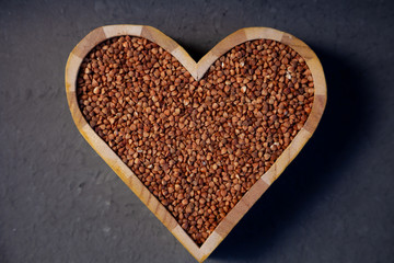 Buckwheat in a wooden plate in the shape of a heart isolated on background. Love for buckwheat porridge. Buckwheat groats, seeds. Useful gluten-free cereal. Close-up, top view.