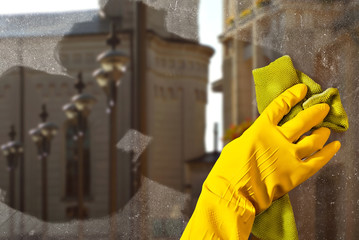 Hand in yellow rubber glove close up. The cleaner washes the window with. Dirty dusty glass in sharpness. City street and buildings outside the window. The architecture is blurred in the background.