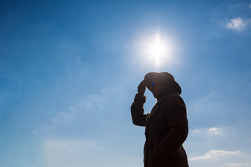 Silhouette of a girl in a hat and coat on a background of the sky with clouds and the sun.
