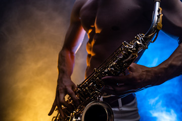 Fototapeta na wymiar Muscular man with naked torso playing on saxophone with smoked colorful background 