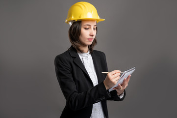 Portrait of wonderful young business woman wearing a protection helmet takes notes, poseing on isolated gray background