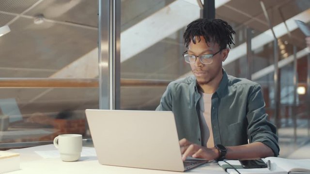 Tracking shot of young African American man with braids typing on laptop while working at desk in modern office