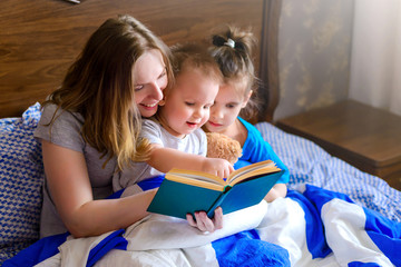 Happy family mother, child daughter, son kid read a book in bed in the morning, before going to bed. Family education. Classes at home during quarantine. Early childhood development. World book day.
