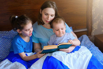 Happy family mother, child daughter, son kid read a book in bed in the morning, before going to bed. Family education. Classes at home during quarantine. Early childhood development. World book day.