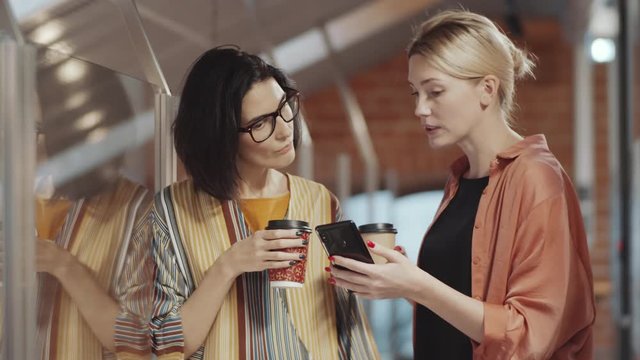 Pretty blonde businesswoman holding disposal coffee cup and discussing something on smartphone screen with brunette female colleague while standing beside glass wall in office