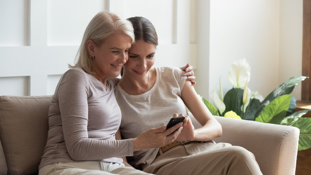 Older Mother And Adult Daughter Hugging, Using Phone Together, Young Woman And Mature Mum, Grandmother Looking At Smartphone Screen, Watching Video Or Photos, Sitting On Cozy Sofa At Home Together