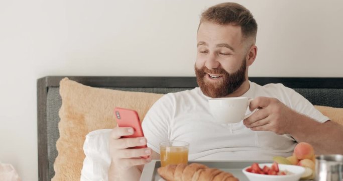 Side view of young man making amazed facial expession while using his modern mobilephone.Guy in 30s having breakfast and drinking coffee while getting shocking news. Concept of tech and life.