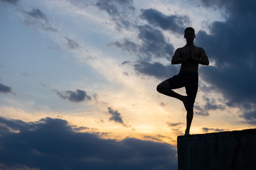 Fit young man practices sun salutation yoga on the edge of cliff at sunset