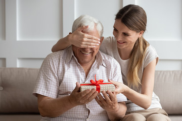 Smiling adult daughter closing older father eyes, preparing surprise, holding gift box with red bow in hand, young woman congratulating dad with birthday or fathers day, sitting on couch at home