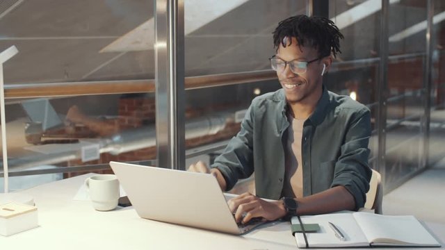 Cheerful African American Man Putting In Wireless Earphones, Talking And Waving At Front Camera Of Laptop While Web Conferencing From Office