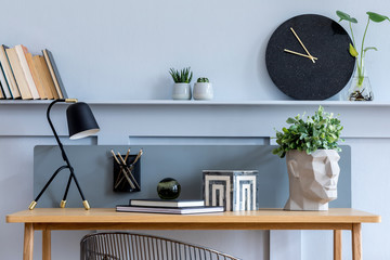 Stylish scandinavian living room interior with wooden desk, chair, wood panleing with shelf, table lamp, plants, black clock, office supplies and elegant accessories in modern home decor.