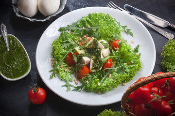 kitchen table with fresh mozzarella salad, lettuce and arugula leaves, pesto sauce, fresh cherry tomatoes in a basket
