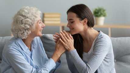 Happy millennial girl sit on couch cuddle caress mature mom enjoy close intimate time together, smiling elderly mother and grown-up adult daughter hold hands spend weekend at home together