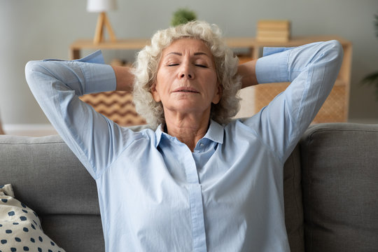 Calm old woman sit relax on cozy couch in living room daydream or take nap at home, relaxed mature female rest on sofa with eyes closed, enjoy peaceful day, breathe fresh air, stress free concept