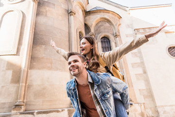 Low angle view of man piggybacking girlfriend with open arms near building in city