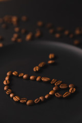 A cup of coffee. The cup on a matte black plate on a black background. Coffee beans around the plate. Black composition. Dark mode style. Coffee time. Closeup. Vertical picture