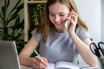 Distance learning online education. A schoolgirl is studying at a computer at home and doing school homework. The girl is talking on the phone and writes.