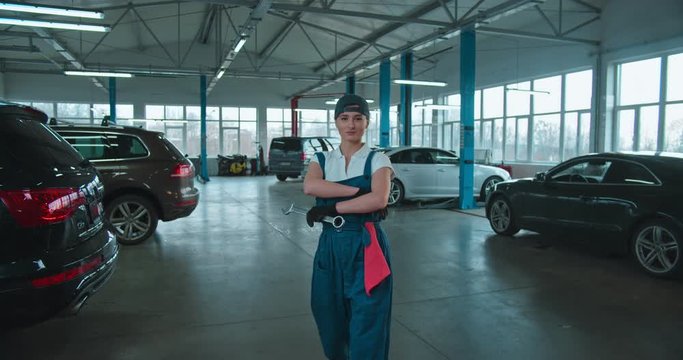 Caucasian young cool attractive woman cars repairment worker in uniform, goggles, hat and with wrench coming close to camera in big garage. Portrait of pretty happy girl mechanic smiling.